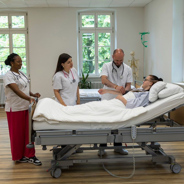 Zwei Pflegeschülerinnen und ein Pflegeschüler stehen im Praxisraum der Pflegeschule an einem Krankenhausbett. Darin liegt eine Patientin. Ihr misst der männliche Azubi den Blutdruck.