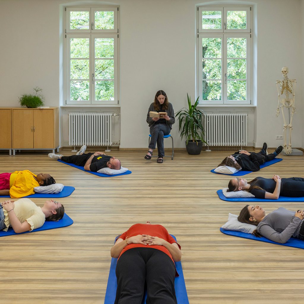 Im Praxisraum der Pflegeschule lieft eine Gruppe von Schülern entspannt auf Yogamatten im Kreis auf dem Boden. Eine Pflegelehrerin sitz auf einem Stuhl und ließt in einem Buch.