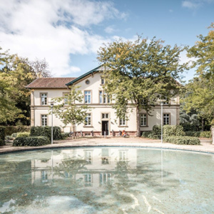 Im Vordergrund ist ein Brunnen, im Hintergrund ein zweistöckiges, älteres aber sehr gepflegtes Gebäude. Drumherum stehen Bäume, die sich im Wasser des Brunnens spiegeln.