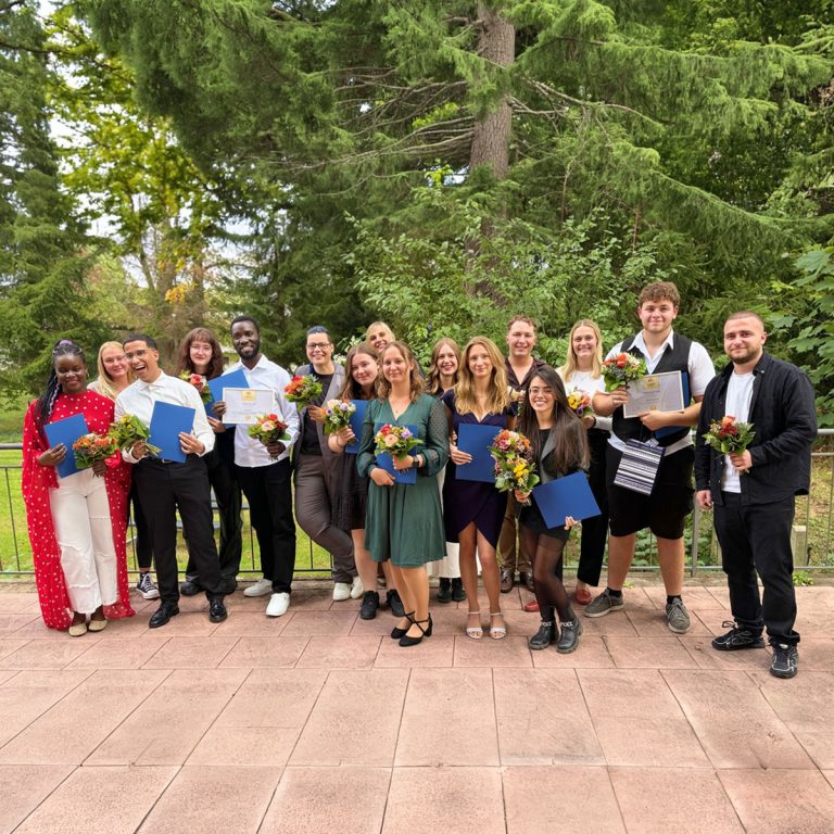 Viele Azubis stehen auf der Terrasse der Schule und halten Geschenke und Blumen in der Hand.